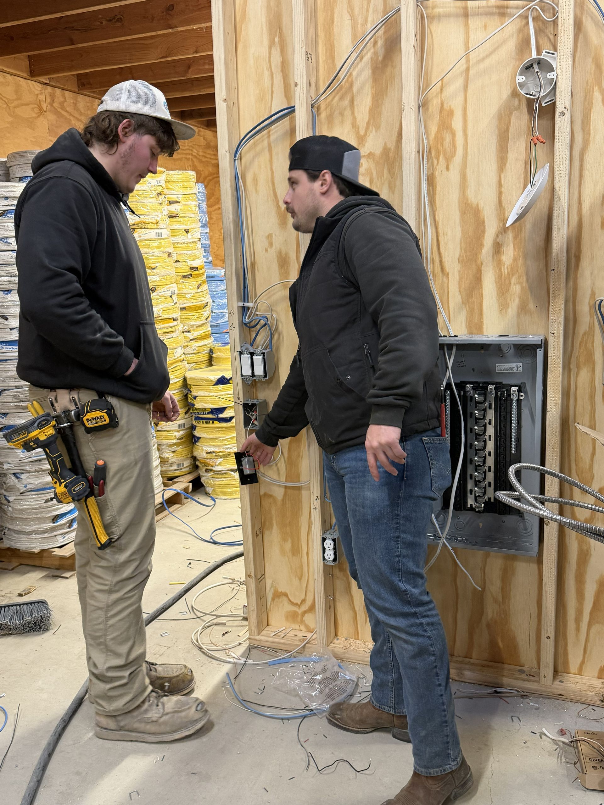 Two construction workers near electrical panel; one points at a switch. Interior with wiring.