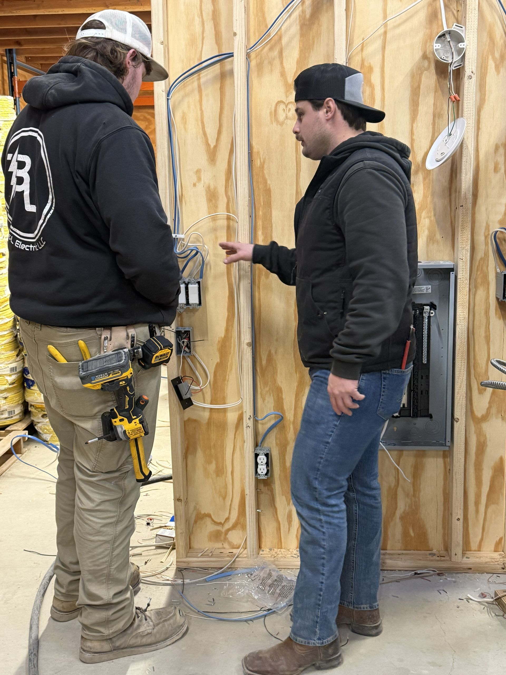 Two electricians inspecting electrical wiring on a wooden wall. One points, the other watches.