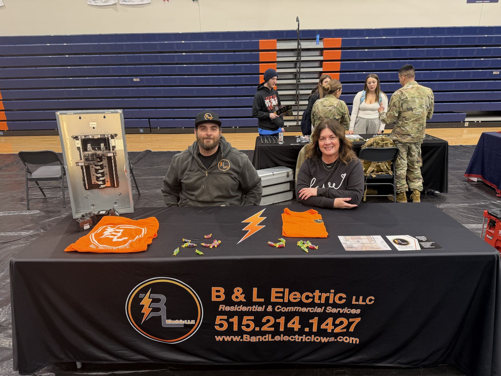 Two people at a B&L Electric booth. A table with company logo, orange accents, and electrical equipment is visible. People mingle in the background.
