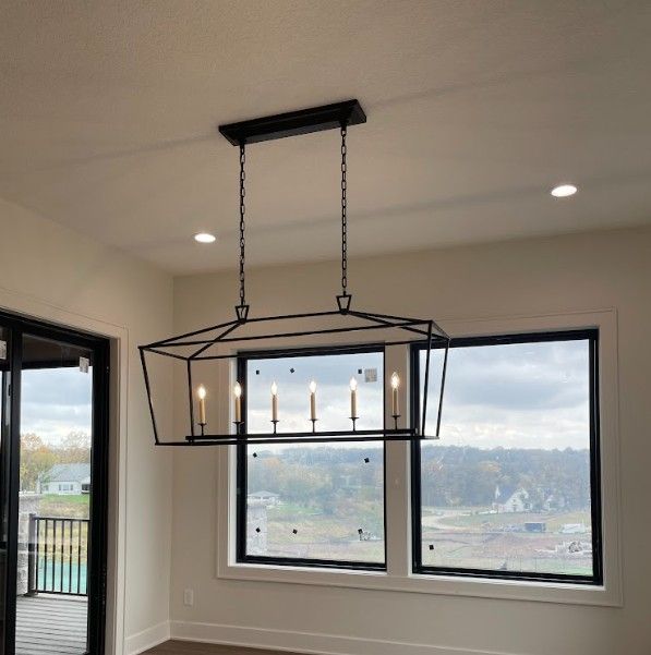 Black geometric chandelier with candle-style lights hangs above a dining area with windows and a view.