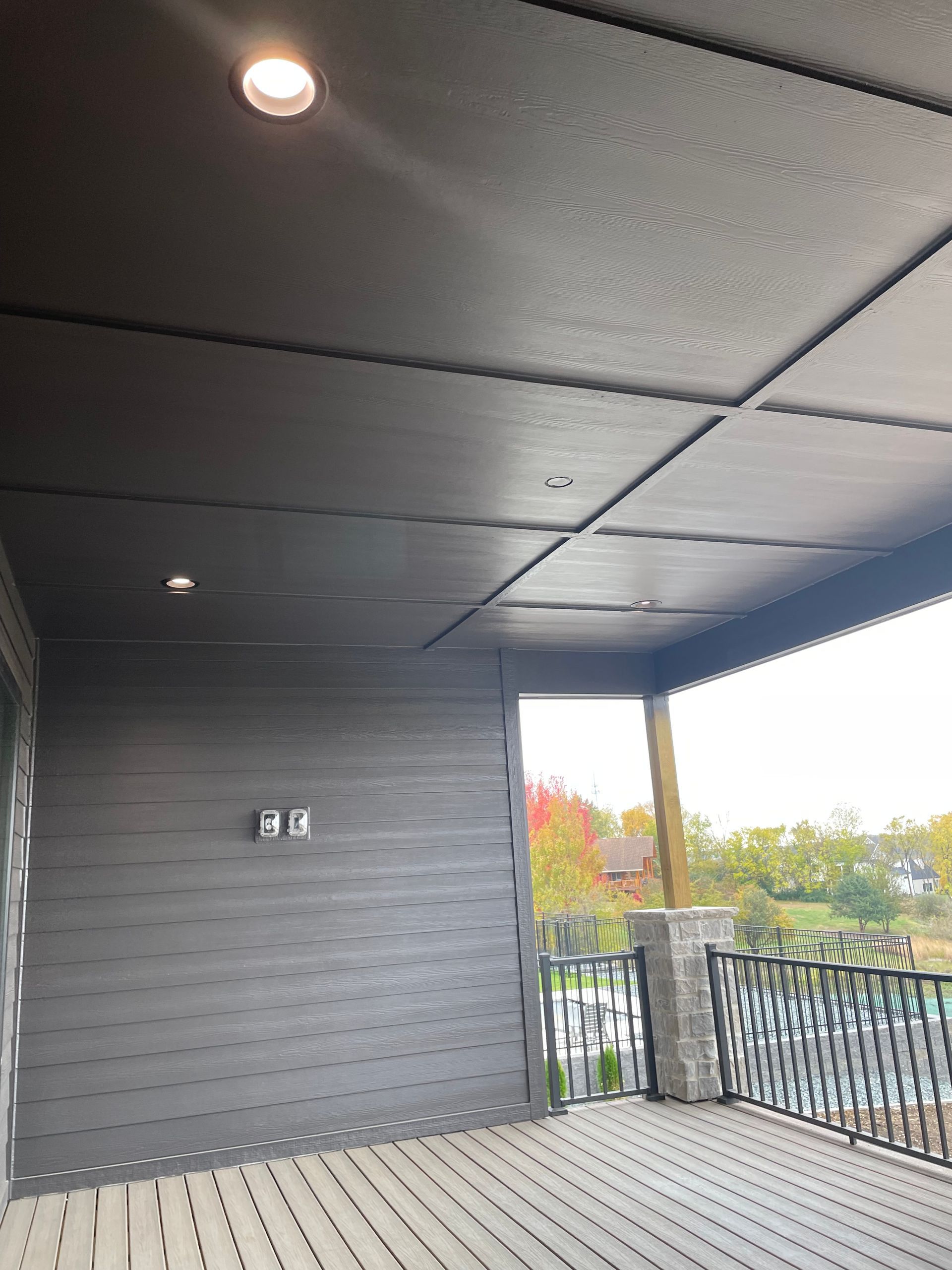 Covered porch with dark gray ceiling, siding, and deck. Recessed lights, stone column, and railing.