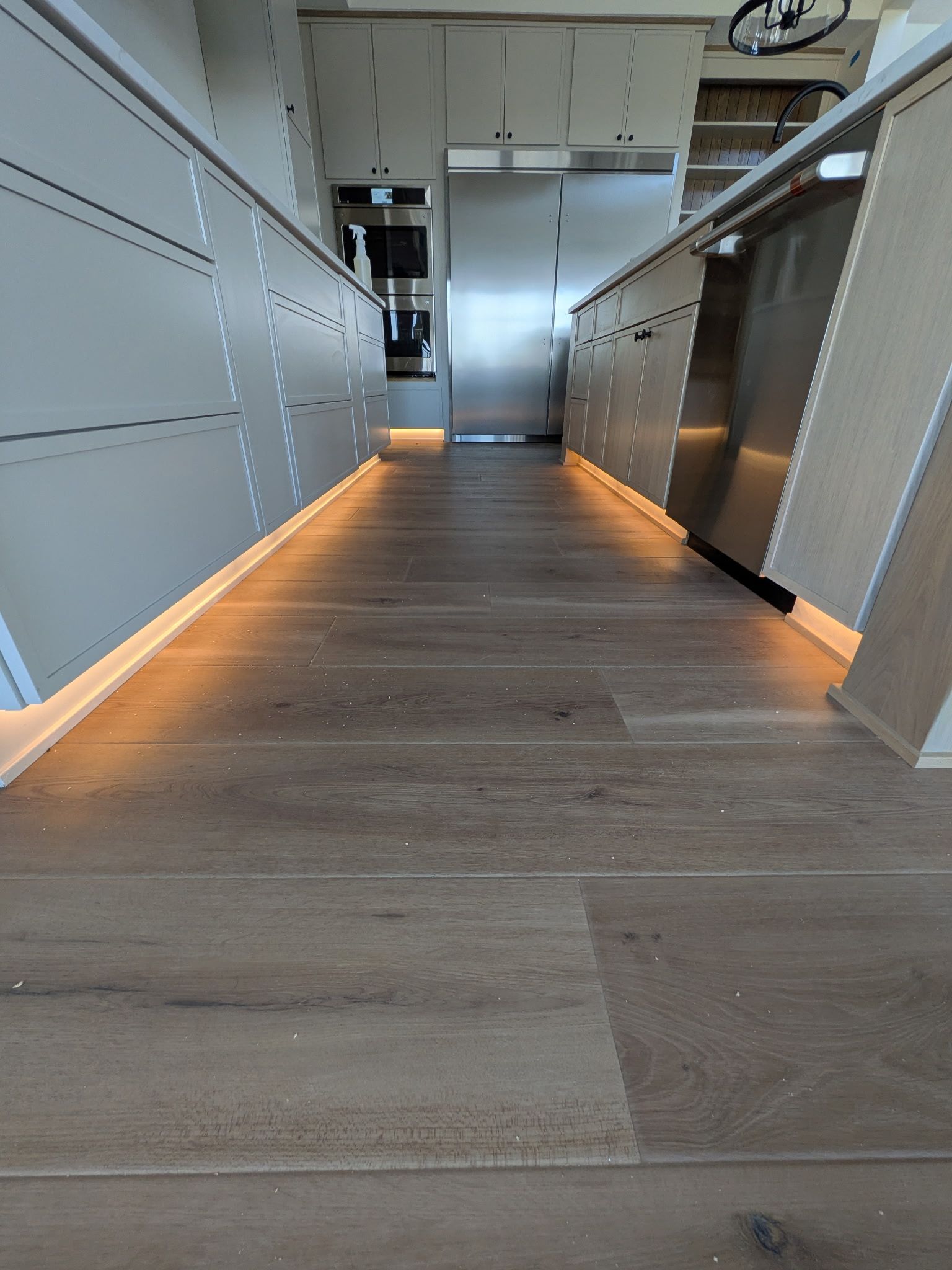 View down a kitchen hallway with a wooden floor and light-colored cabinets, illuminated by strip lighting.