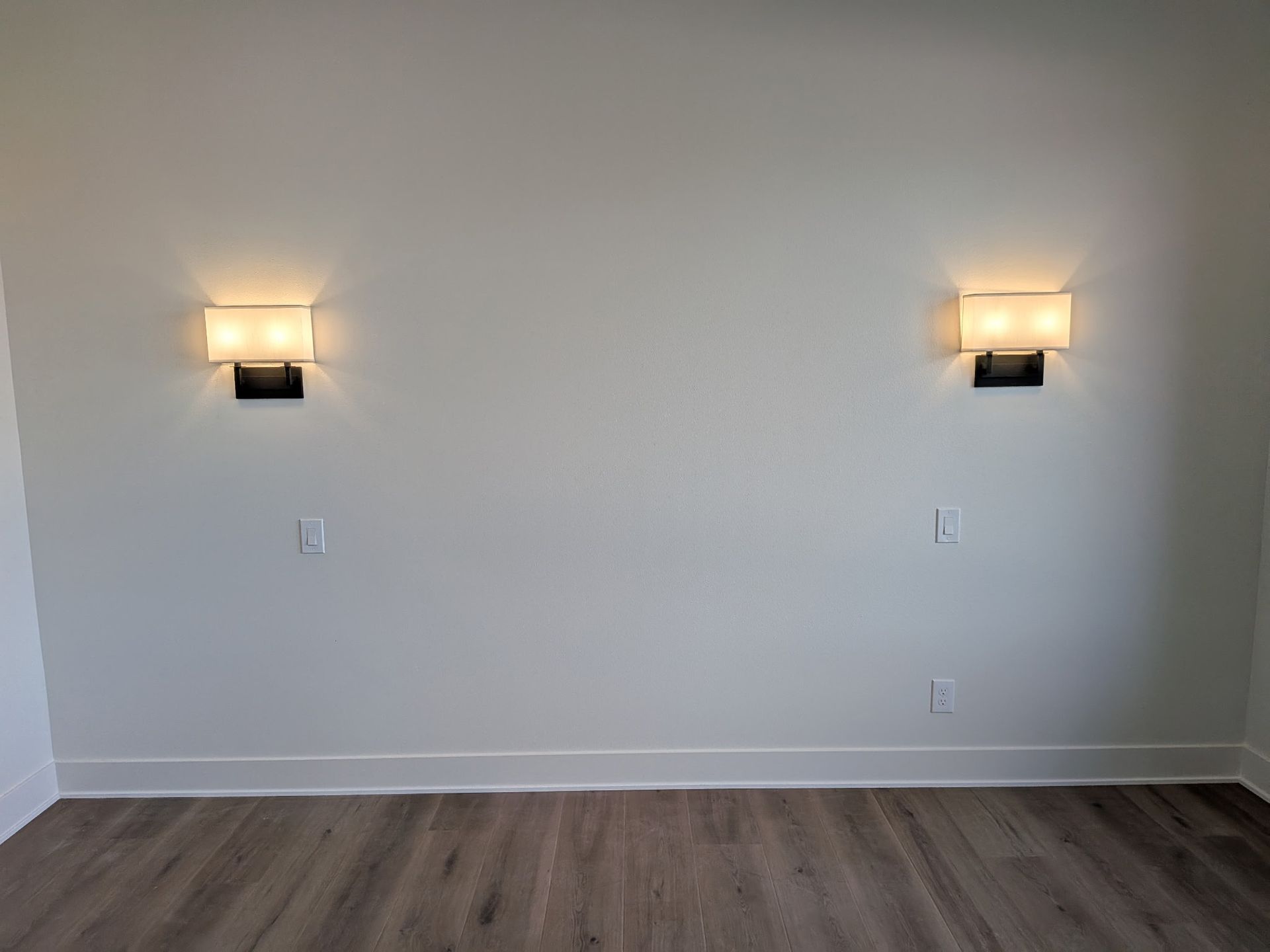Two wall-mounted lights illuminate a white wall with outlets above baseboards and wood-look flooring.
