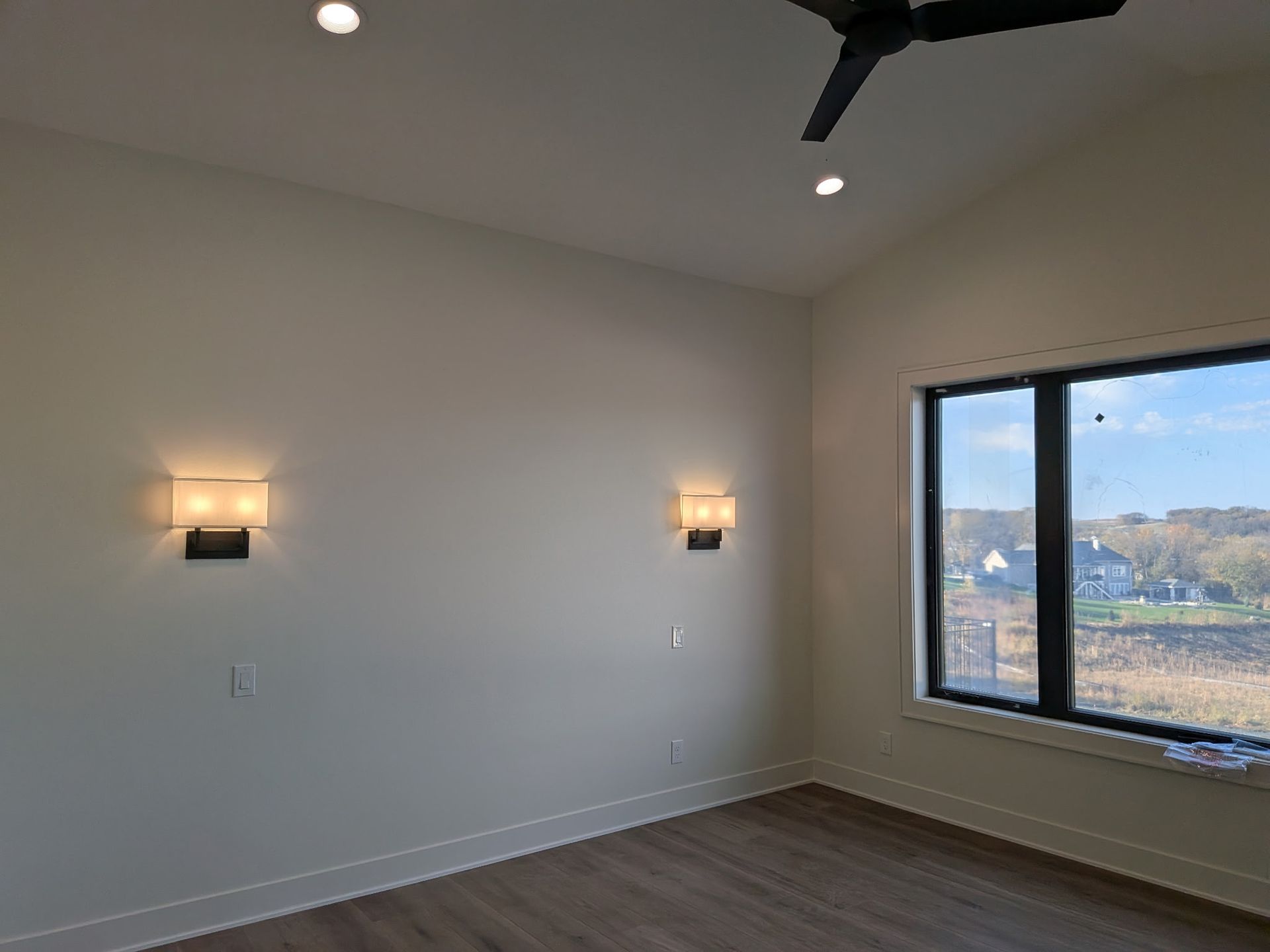 Empty room with white walls, two sconces, a window with a view, ceiling fan, and hardwood floor.