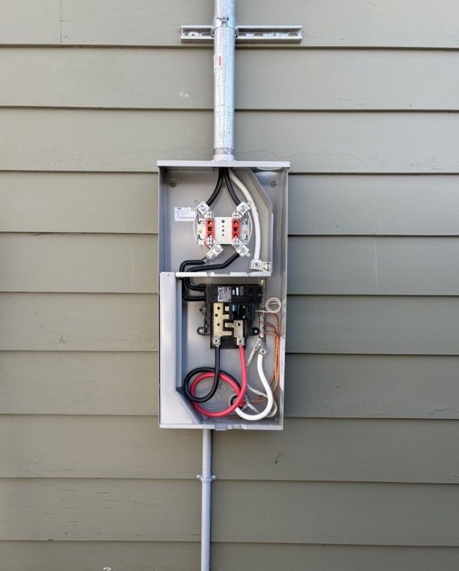 Electrical meter box mounted on a gray siding wall, with exposed wiring and components.