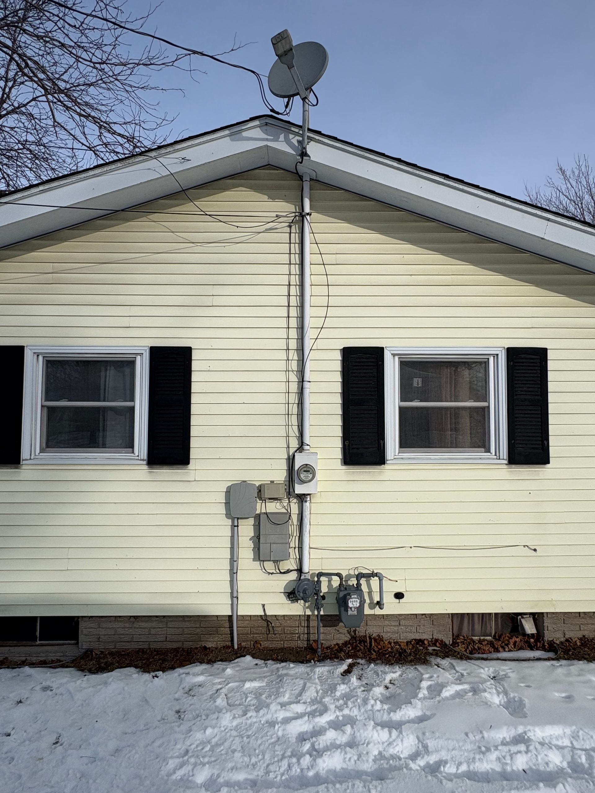 Yellow house with windows, black shutters, and satellite dish on the roof. Gas meter and wiring on the side. Snowy ground.