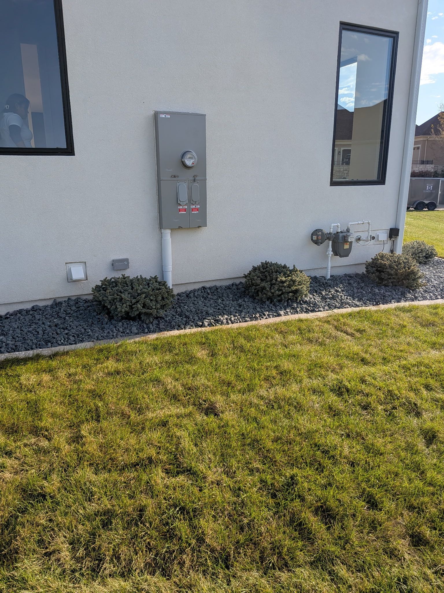 Exterior wall with electrical panel, windows, and bushes against a gravel bed. Green grass in the foreground.