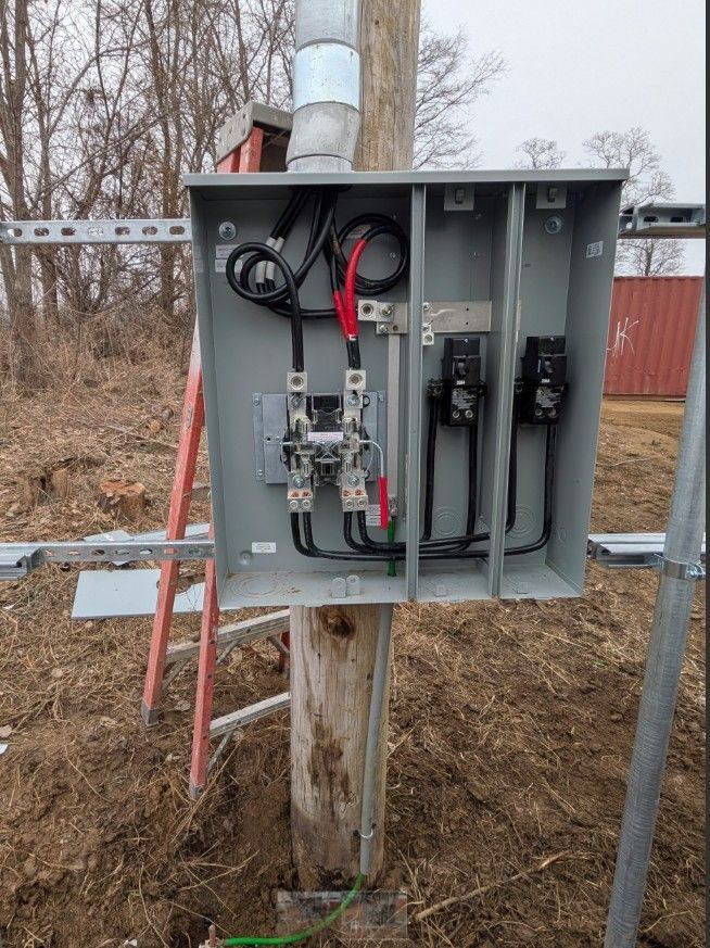 Electrical box mounted on a wooden utility pole with wires, grounding, and ladder visible. Outdoors, daytime. 