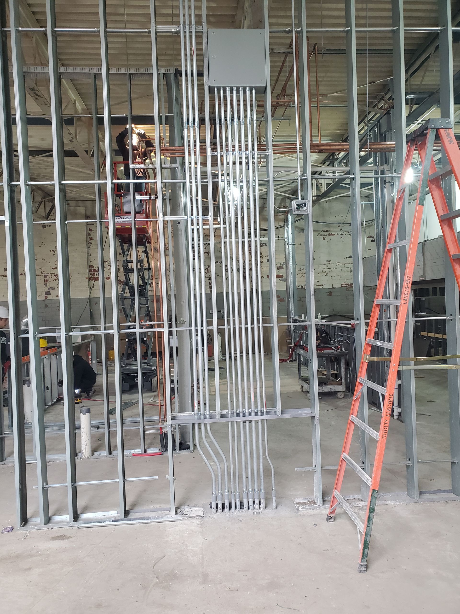 Steel framing with conduit, electrical box, and ladder in a construction area. Workers visible.