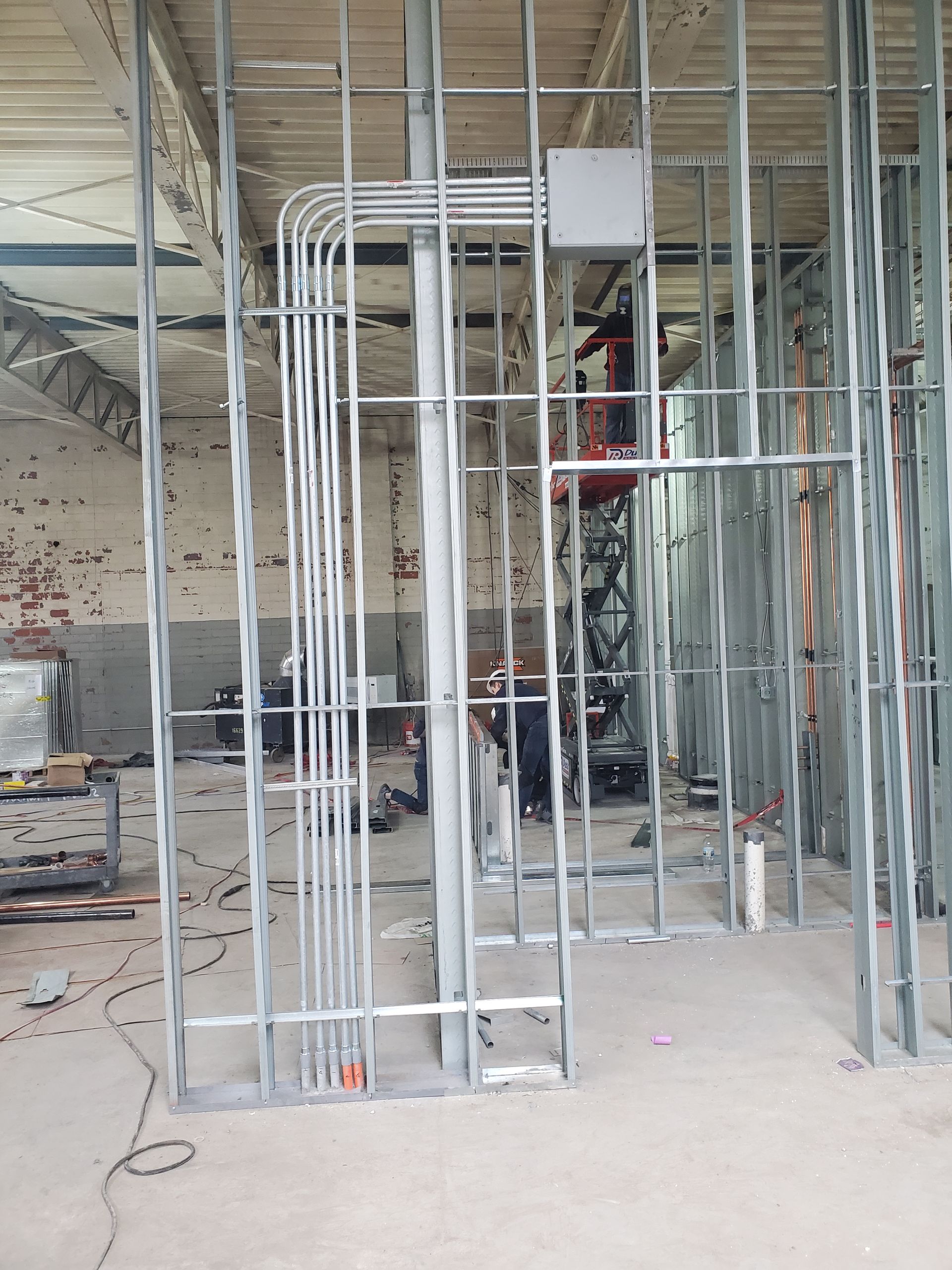 Steel frame construction with electrical conduits and junction box. A scissor lift is visible in the background.