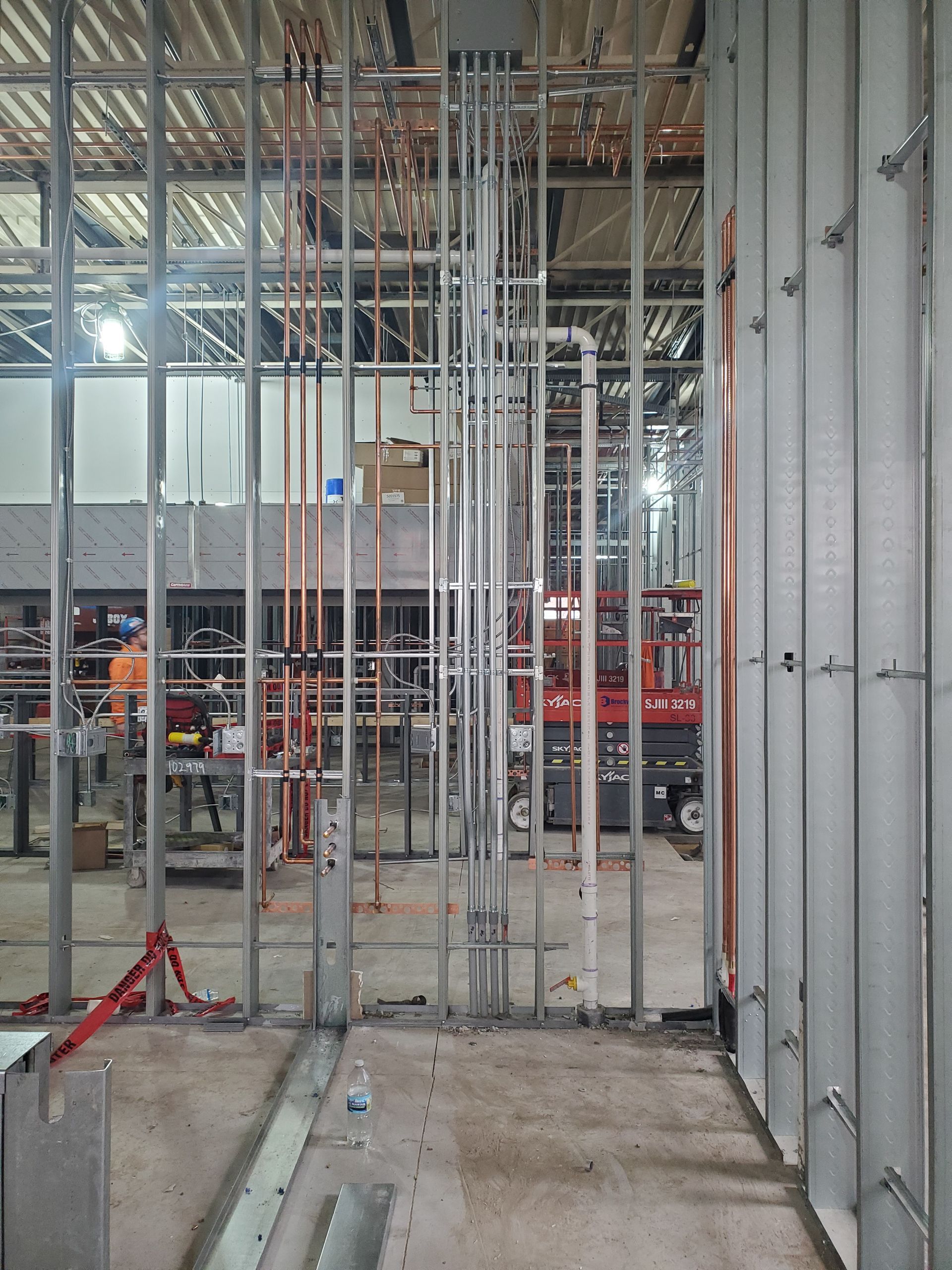 Interior view of a construction site with exposed metal framing, copper pipes, and a red lift in the background.