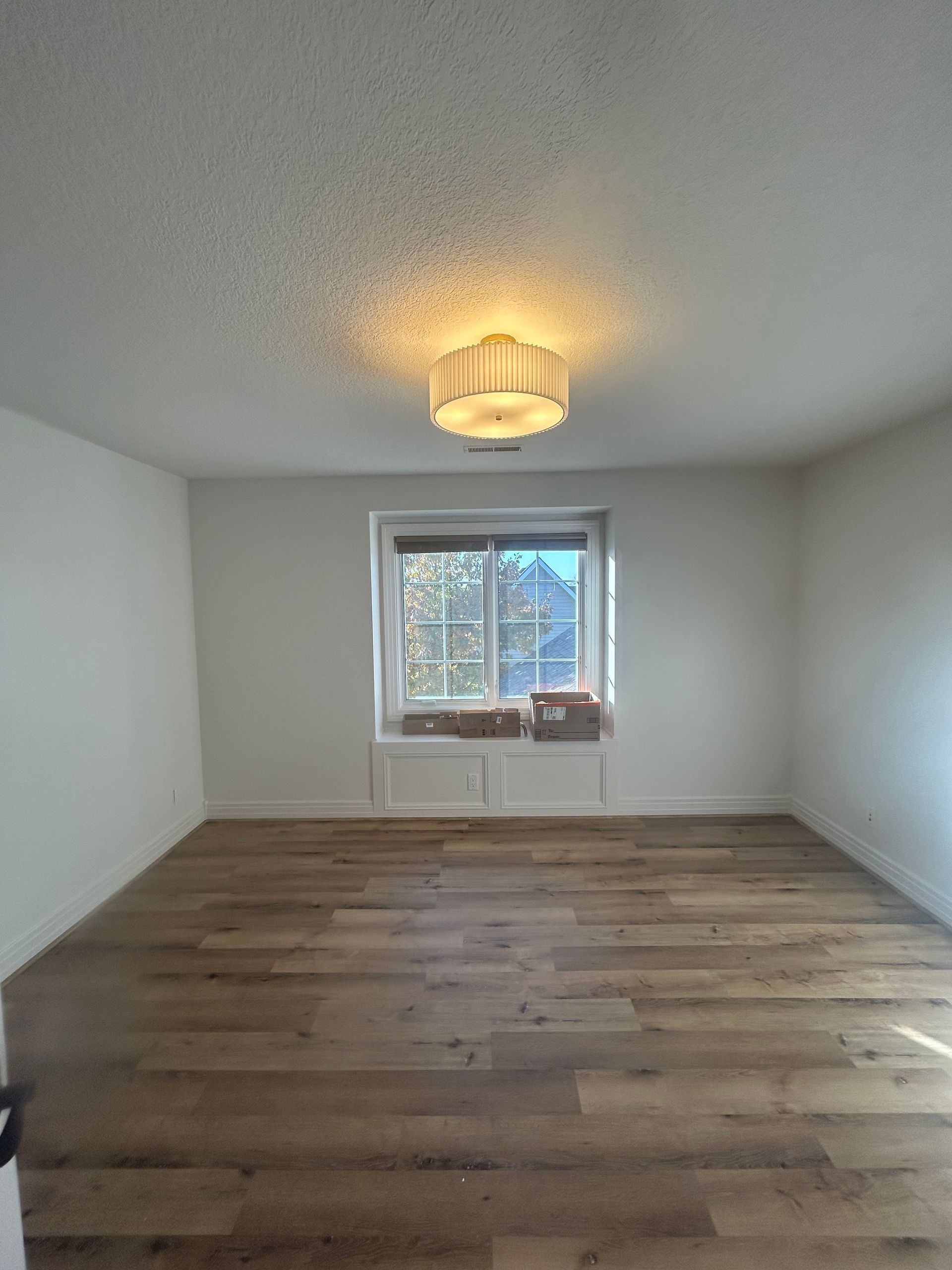 Empty room with wood floors, white walls, and a window centered. Ceiling light fixture.