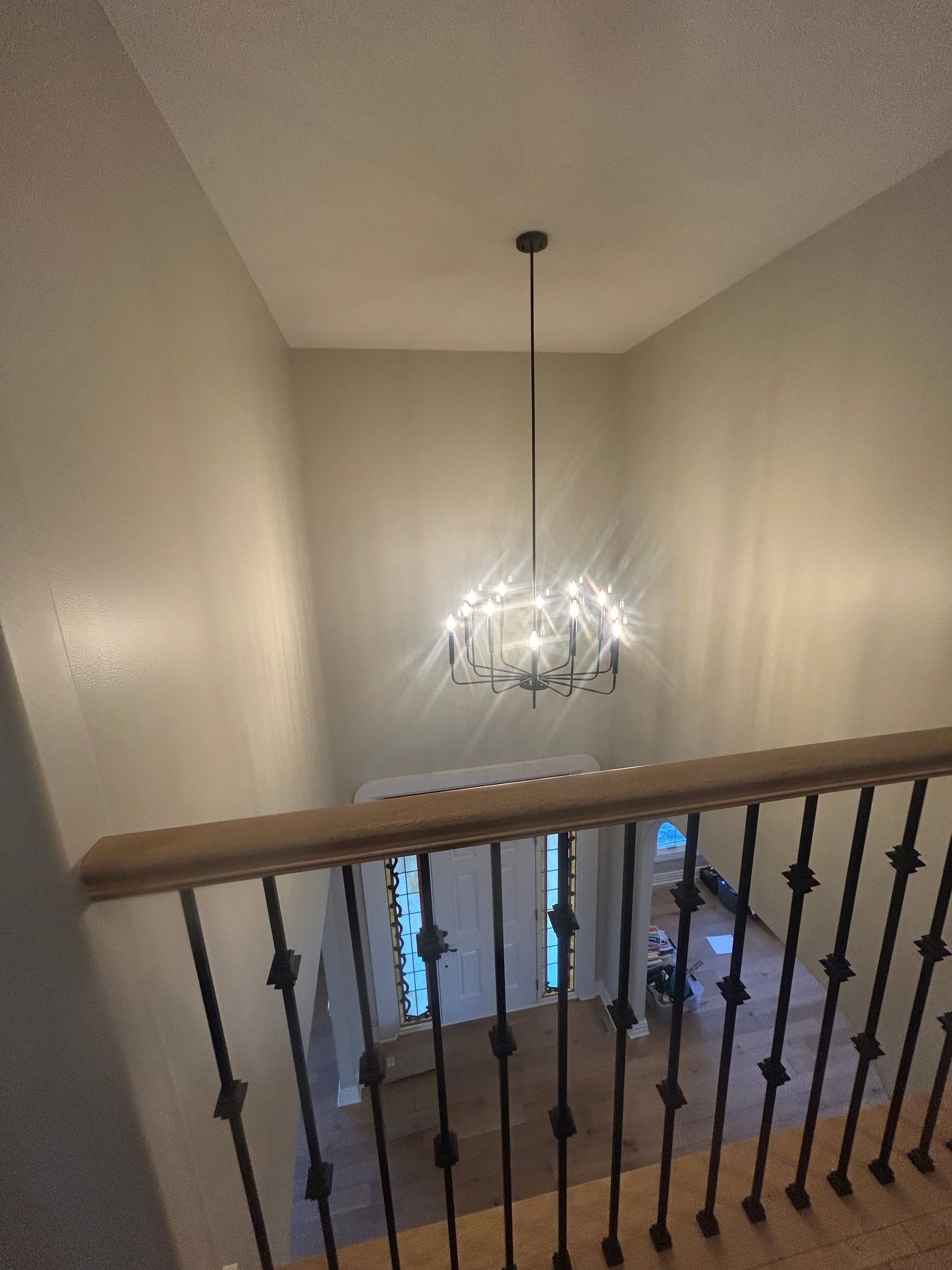 View from a balcony of a foyer with a chandelier hanging. Light beige walls with black railing.
