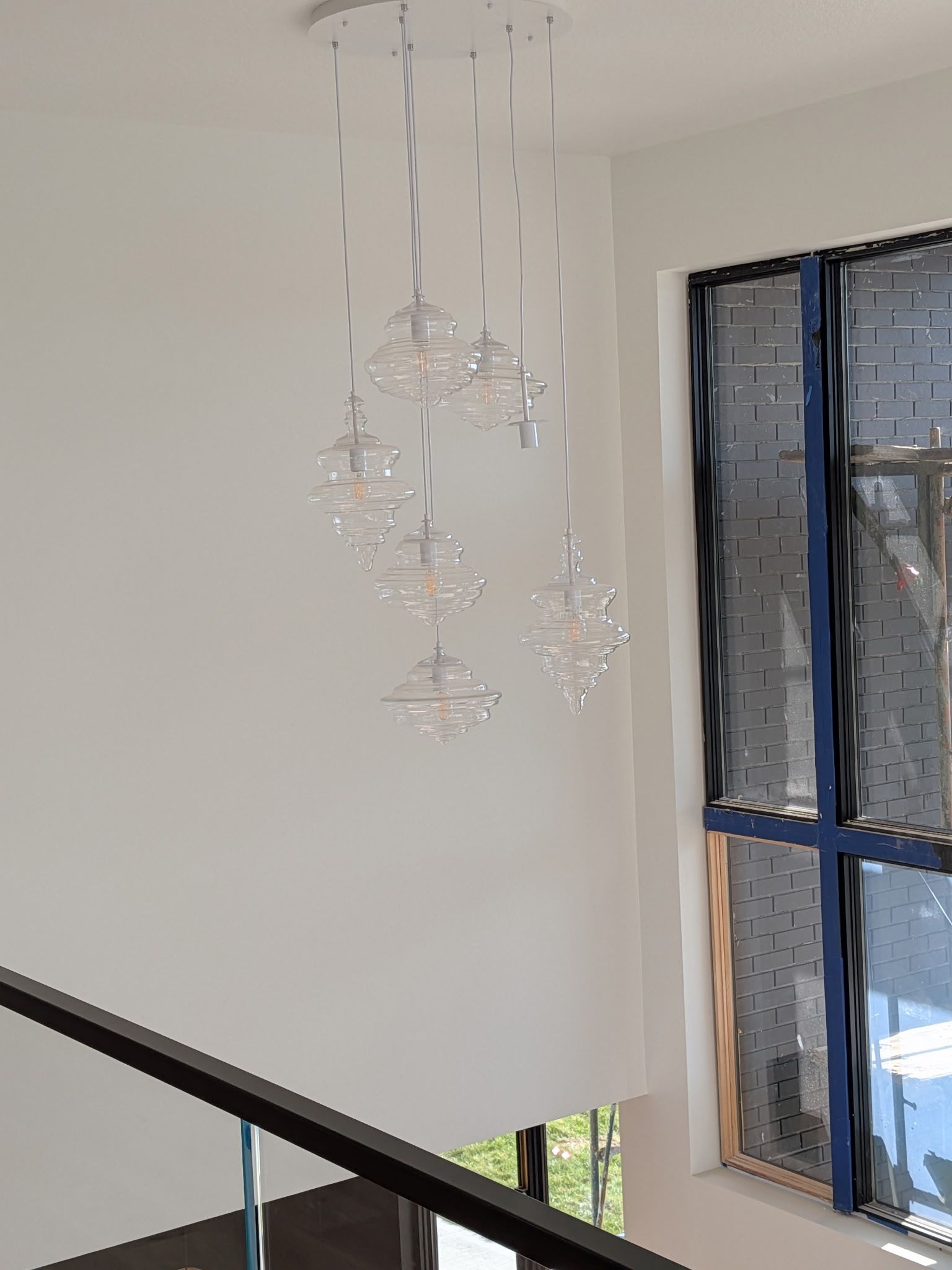 Modern chandelier with crystal orbs hangs in a bright, white-walled stairwell, beside a large window.