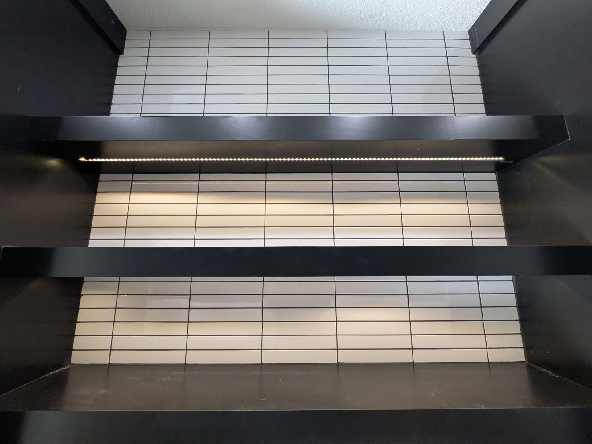 Black shelves with white tiled background, lit by a strip light.