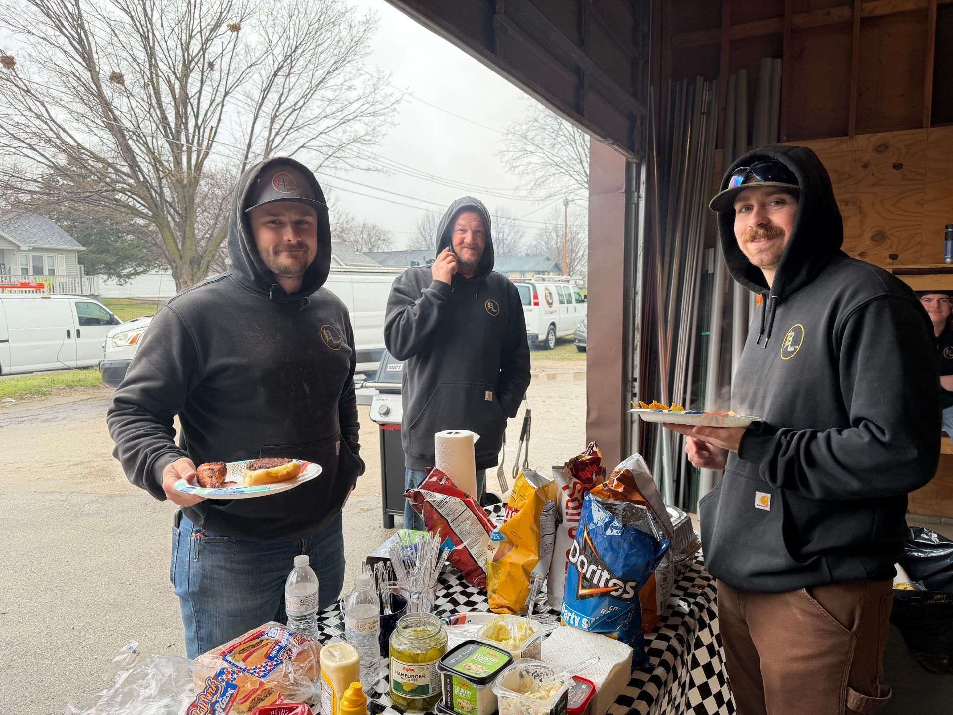 Three hooded people stand by a snack table under a covered outdoor area on a rainy day.
