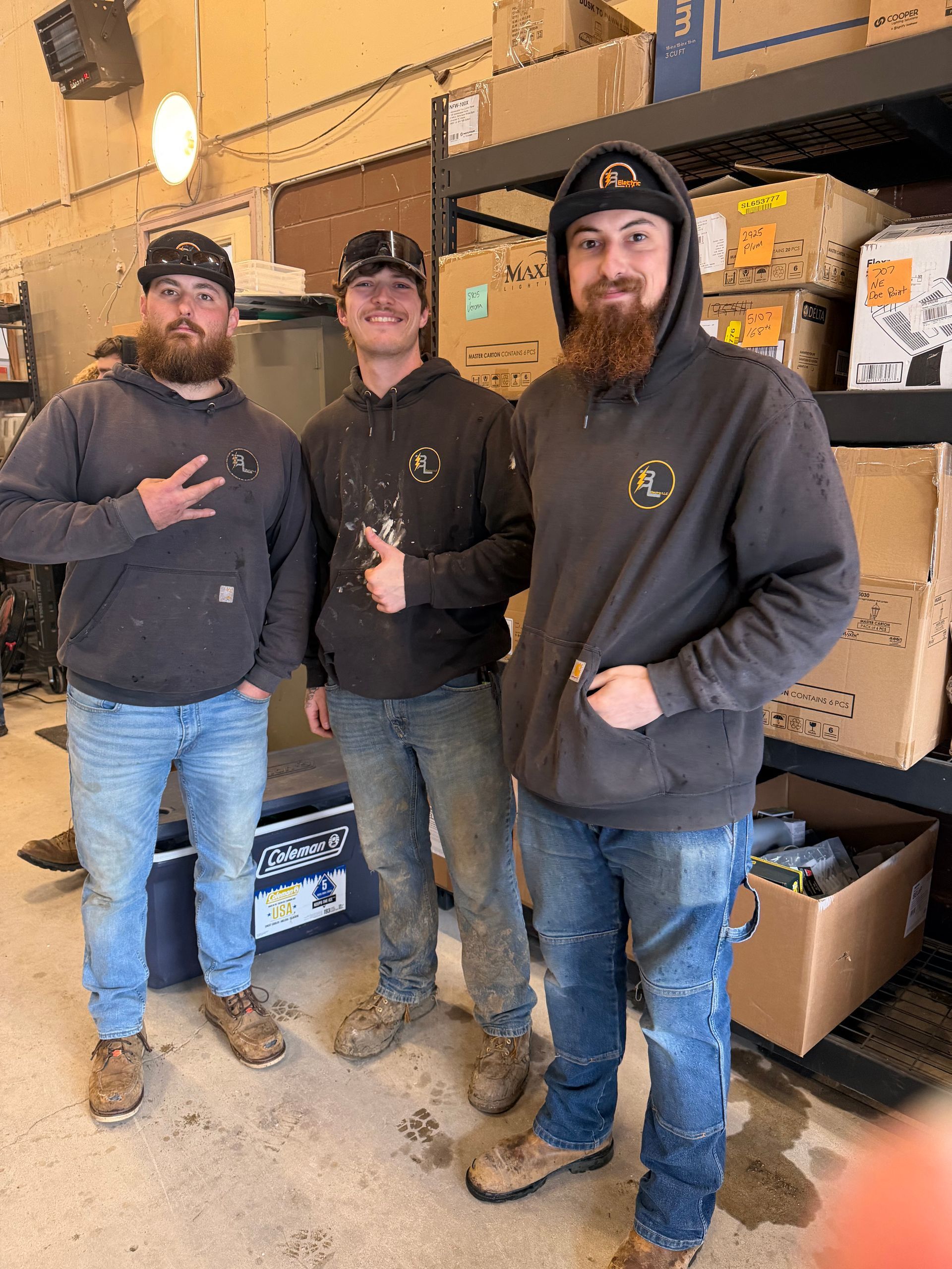 Three workers standing in a warehouse aisle beside stacked boxes, wearing hoodies and boots.