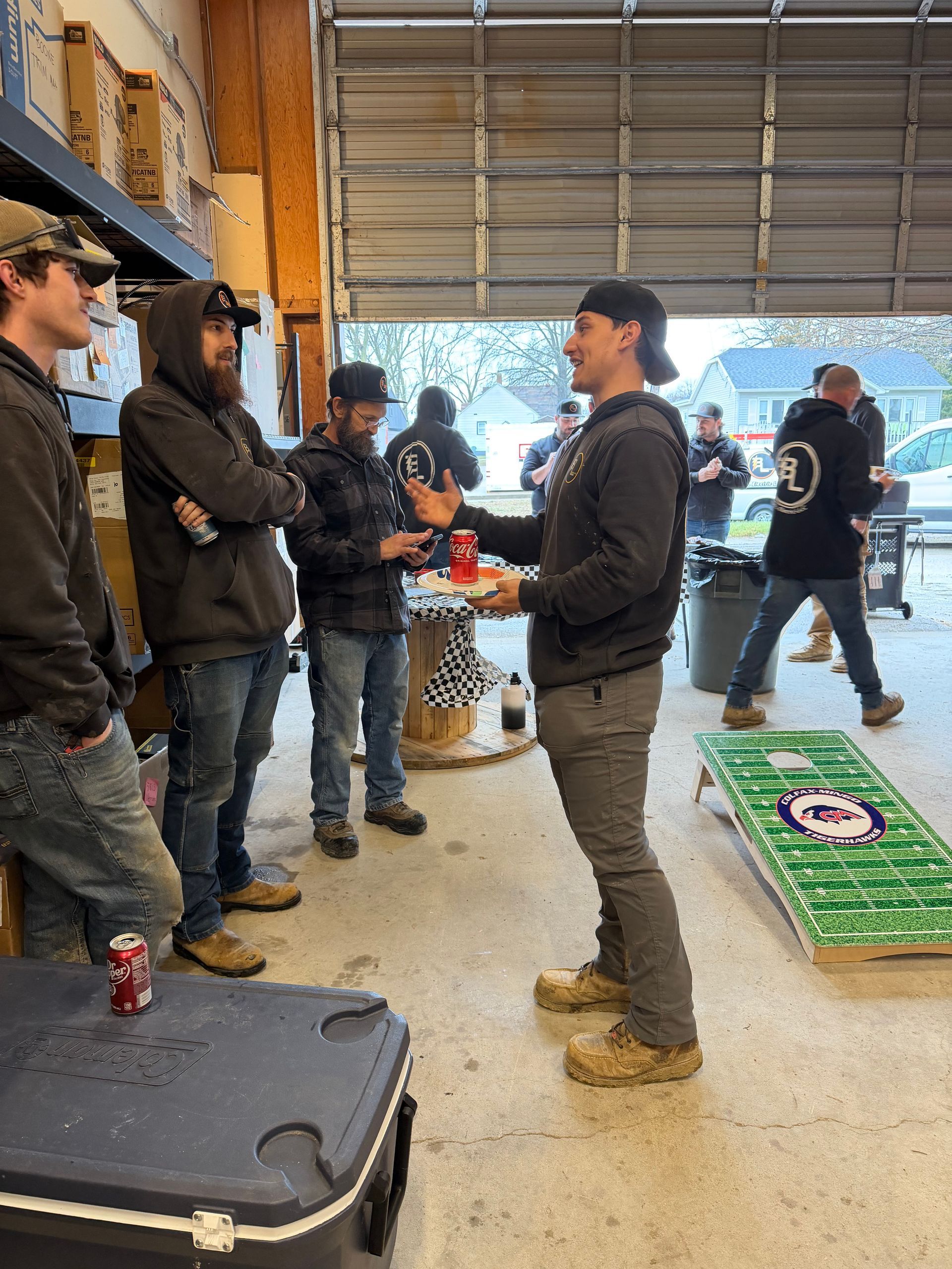 People gathered in a garage-like space, talking near stacked boards and equipment.