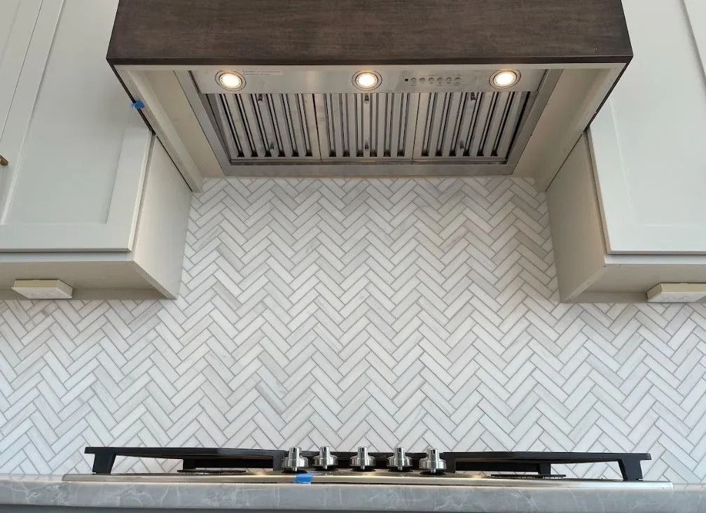 Kitchen with white herringbone tile backsplash, stainless steel range hood, dark wood trim, and gas stovetop.