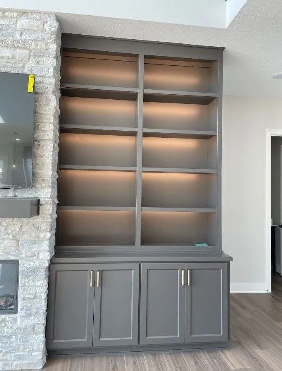 Gray built-in shelving unit with interior lighting and lower cabinet doors, set next to a stone fireplace.