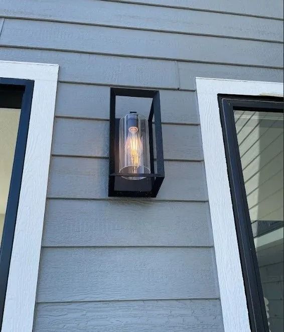 Black outdoor wall light fixture on gray siding between two white-framed windows.