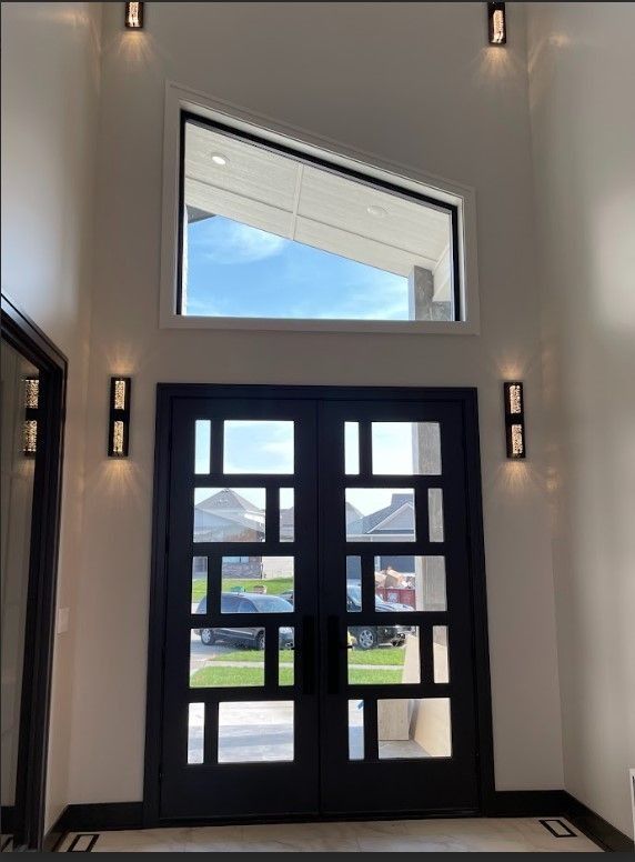 Black geometric double doors with sidelights, overhead window, and wall sconces on a white interior.