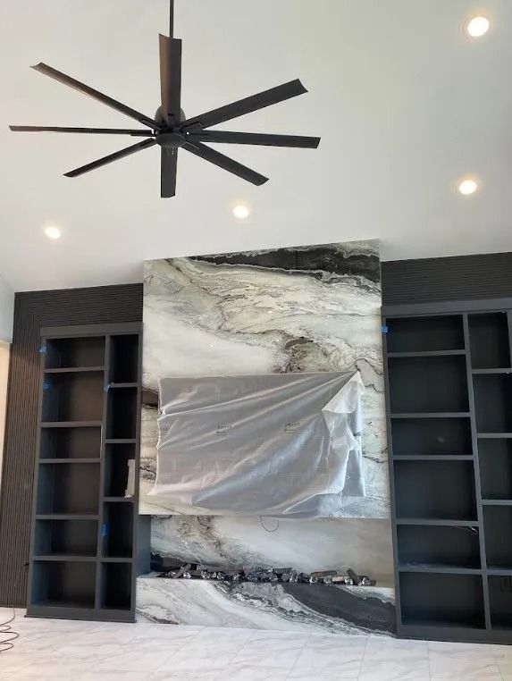 Living room with built-in bookshelves, a stone fireplace, and a ceiling fan. TV is covered.