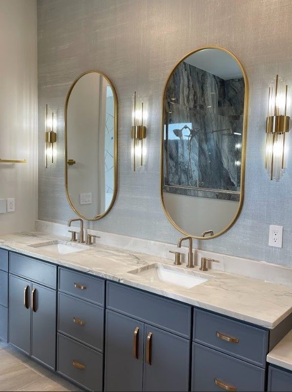 Double vanity bathroom with gray cabinets, marble countertop, gold-framed oval mirrors, and sconces.