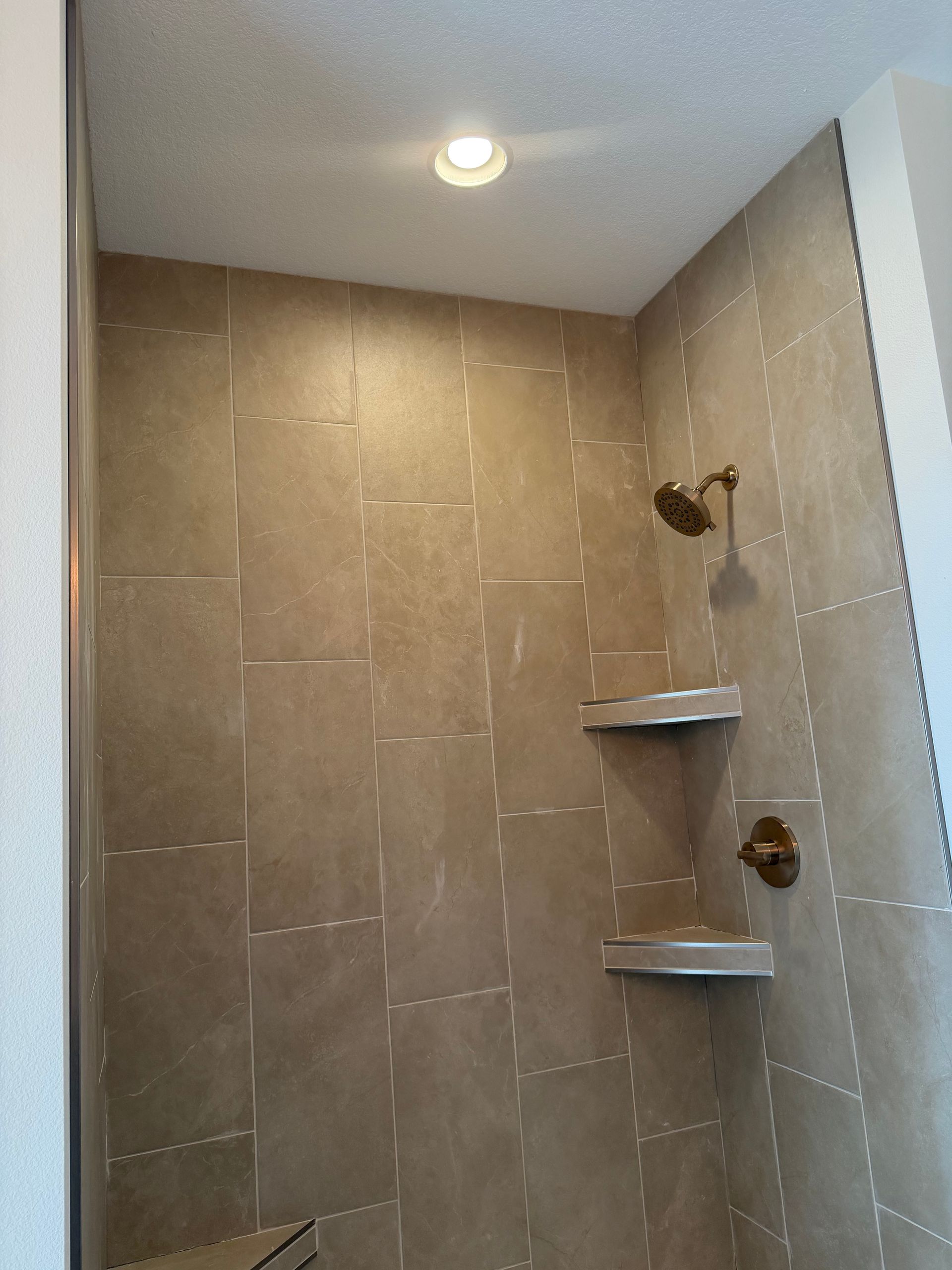 Tiled shower stall with two corner shelves, showerheads, and a ceiling light above.