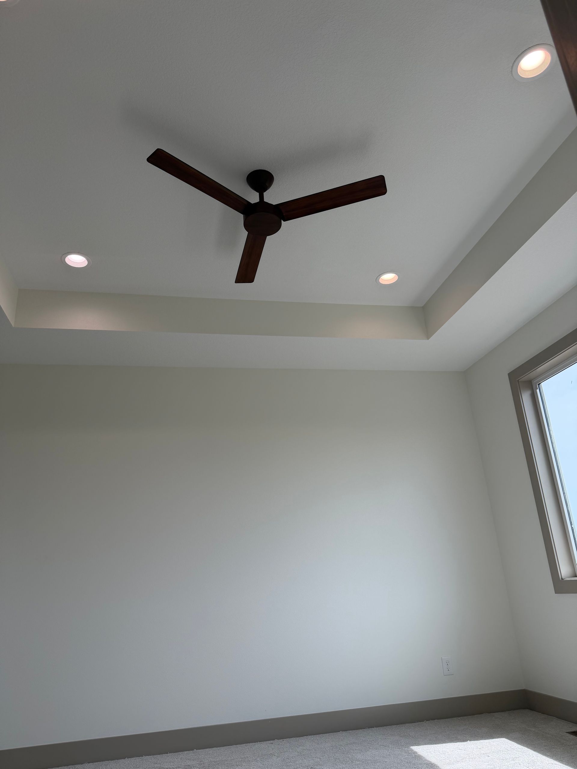Empty white room with tray ceiling, dark ceiling fan, recessed lights, and a window
