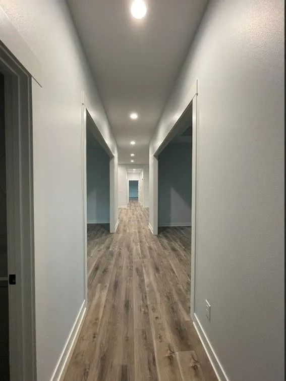 Long, narrow hallway with wood flooring, white walls, and recessed lighting.