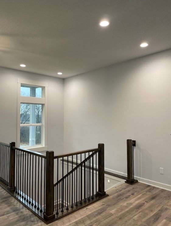 Interior view of a landing with a dark wood railing, window, and recessed lighting. Light gray walls and wood-look flooring.