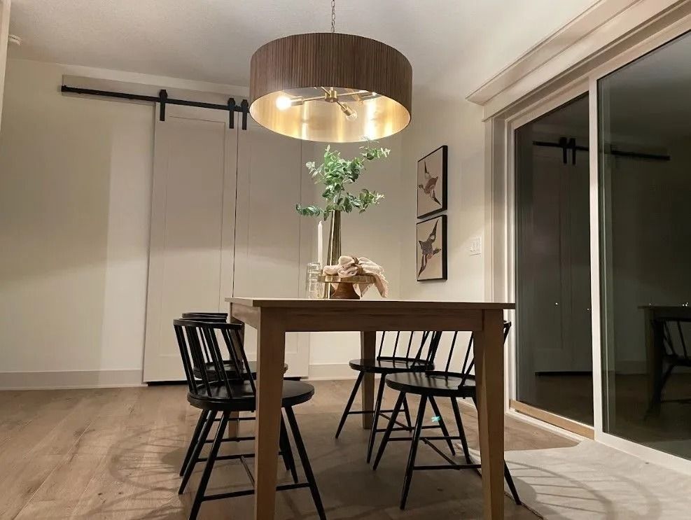 Dining room with wooden table, black chairs, and a round pendant light. Sliding glass door and white walls.