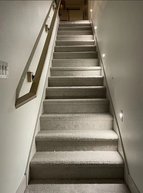 Staircase with carpeted steps and a wooden handrail, lit by small lights on the wall.