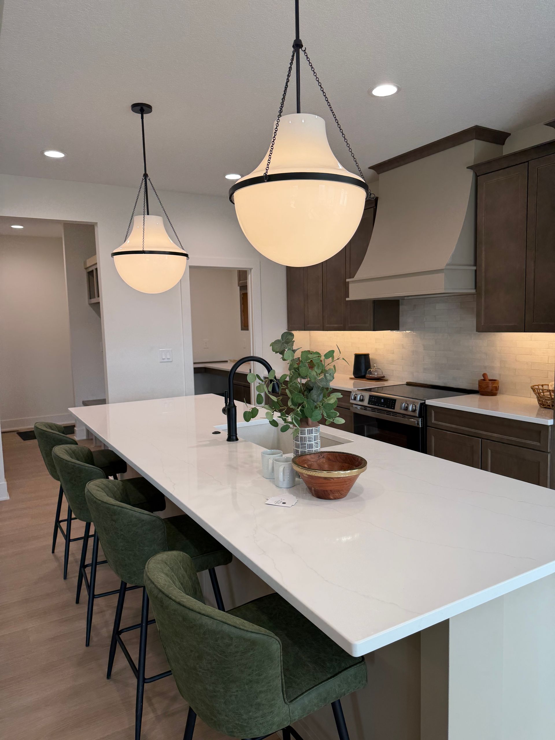 Modern kitchen with white island, green chairs, pendant lights, and dark cabinets.