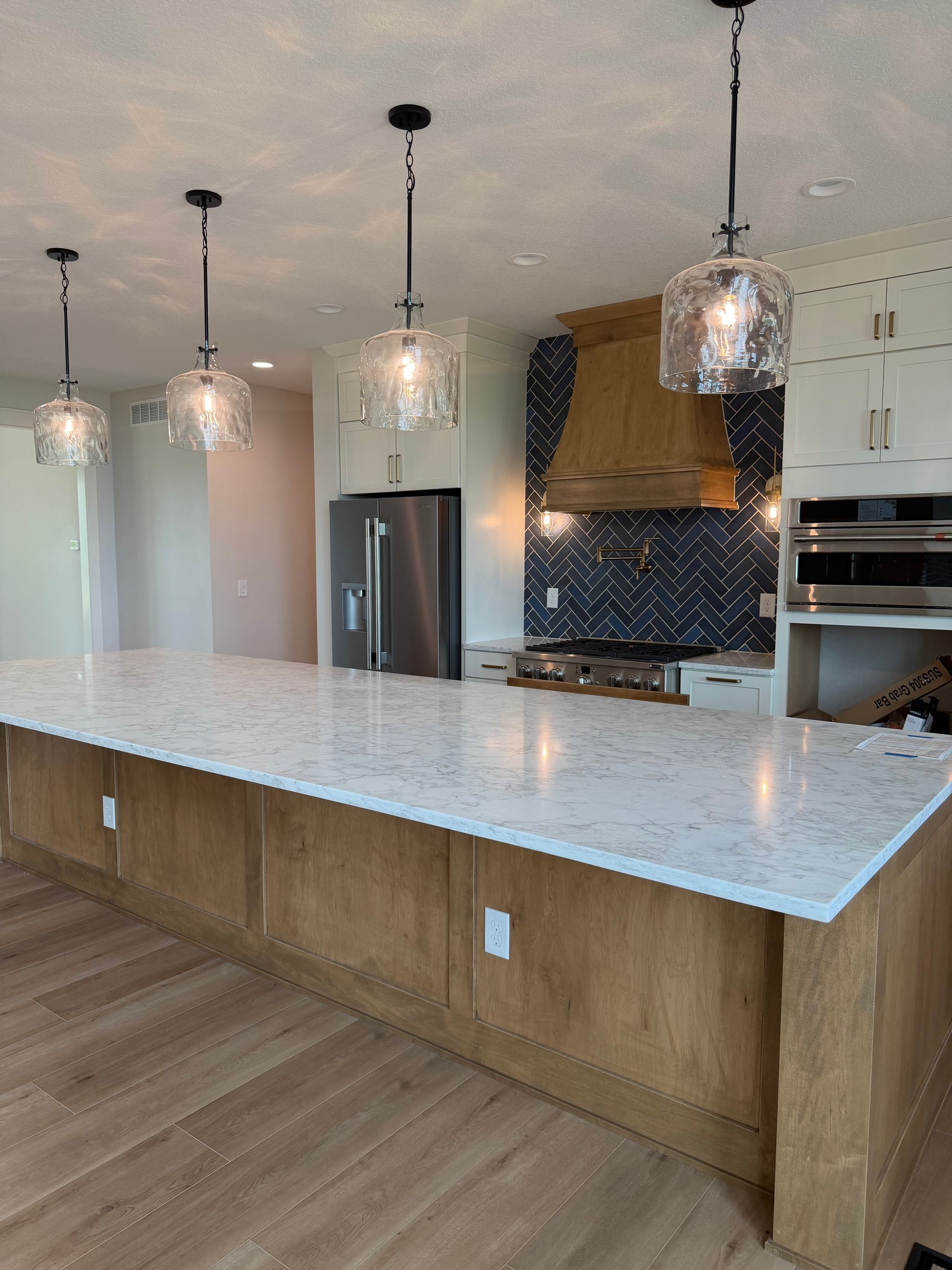 Modern kitchen with a large white island, pendant lights, stainless fridge, and dark herringbone backsplash.