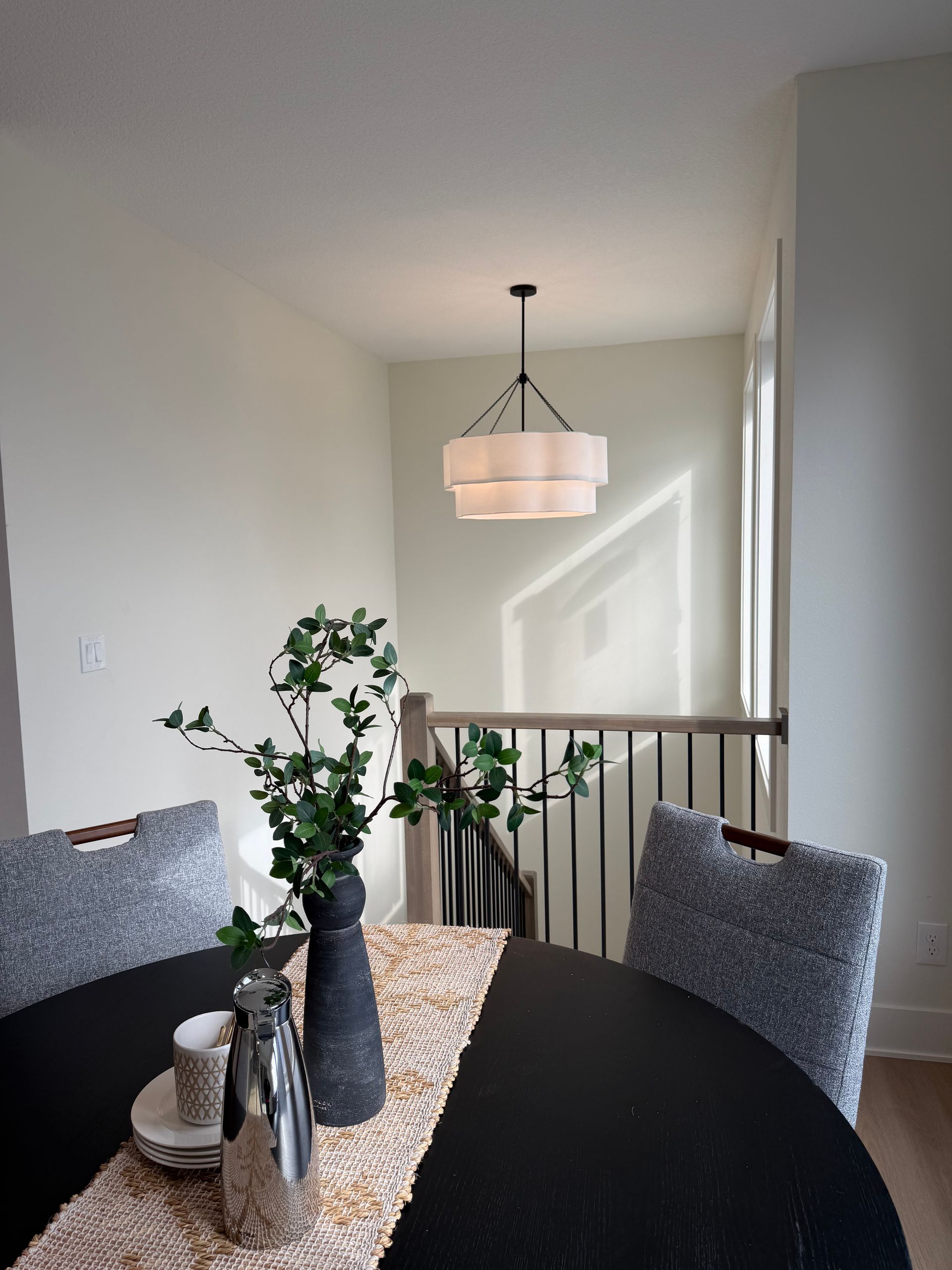 Dining room with a round black table, chairs, and a layered chandelier. A vase with greenery sits on the table.