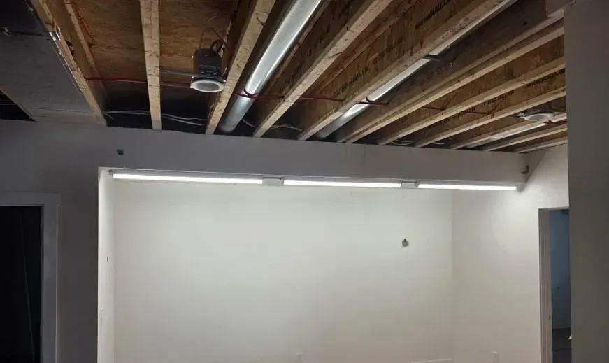 Empty room with exposed wooden ceiling beams, white wall, and fluorescent light fixture