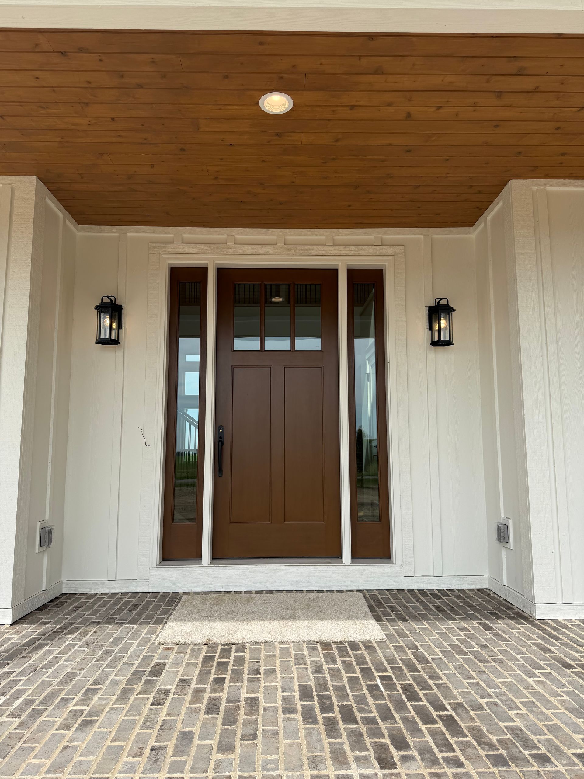 Front entrance with wooden double doors, sidelights, black lanterns, and brick pavers under a covered porch
