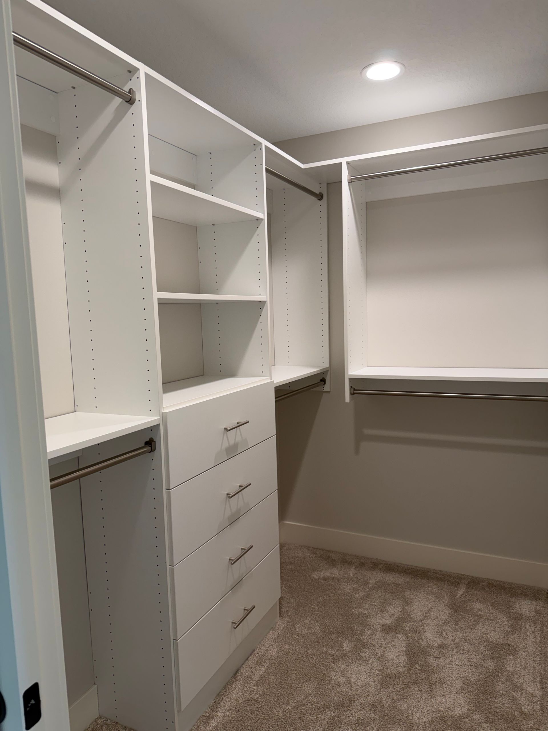 Walk-in closet with white built-in shelves and drawers, beige carpet, and hanging clothes on the right.