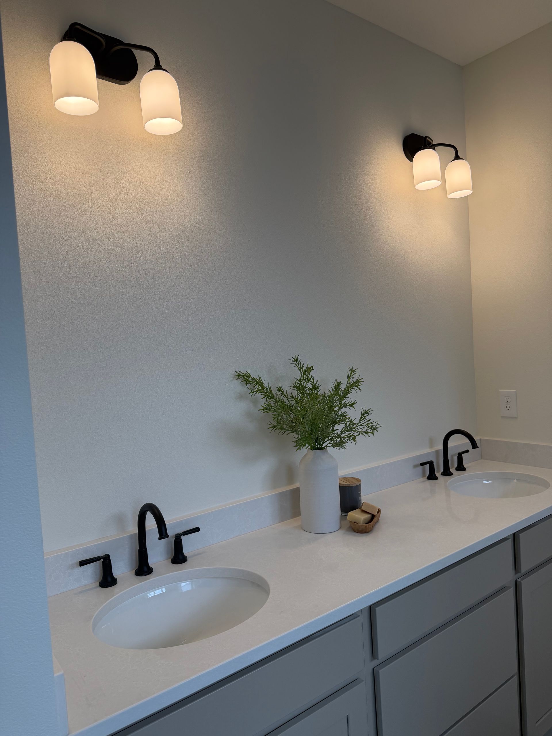 Bathroom with two sinks, gray cabinets, white countertops, black faucets, sconce lights, and a vase with greenery.