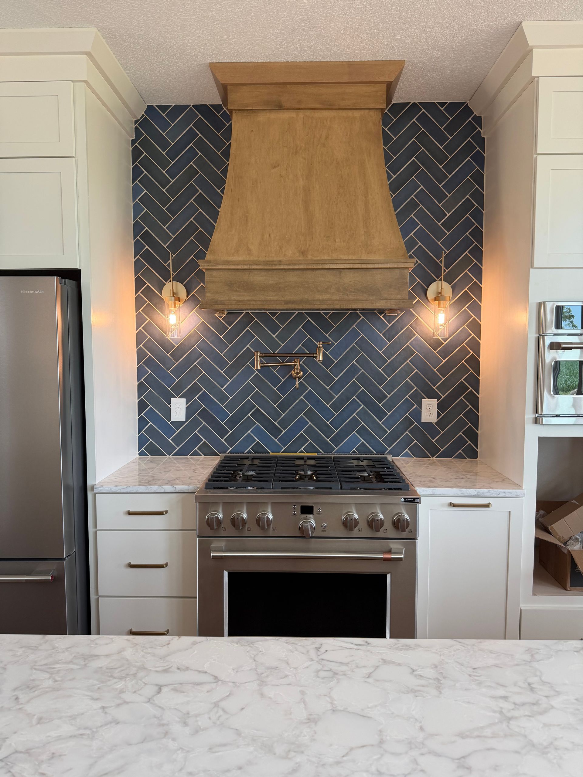 Modern kitchen with blue chevron tile backsplash, gold range hood, white cabinets, and marble island countertop