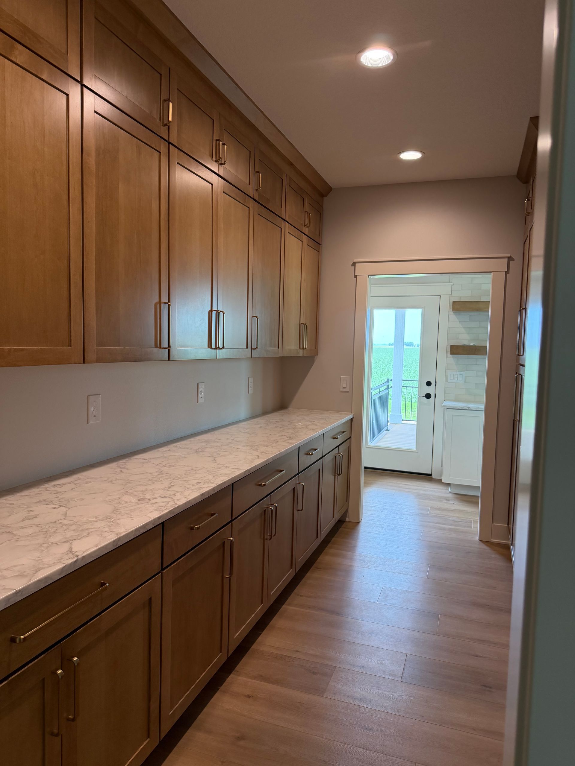 Long kitchen with wood cabinets, light countertops, wood floor, and doorway to a bright entryway