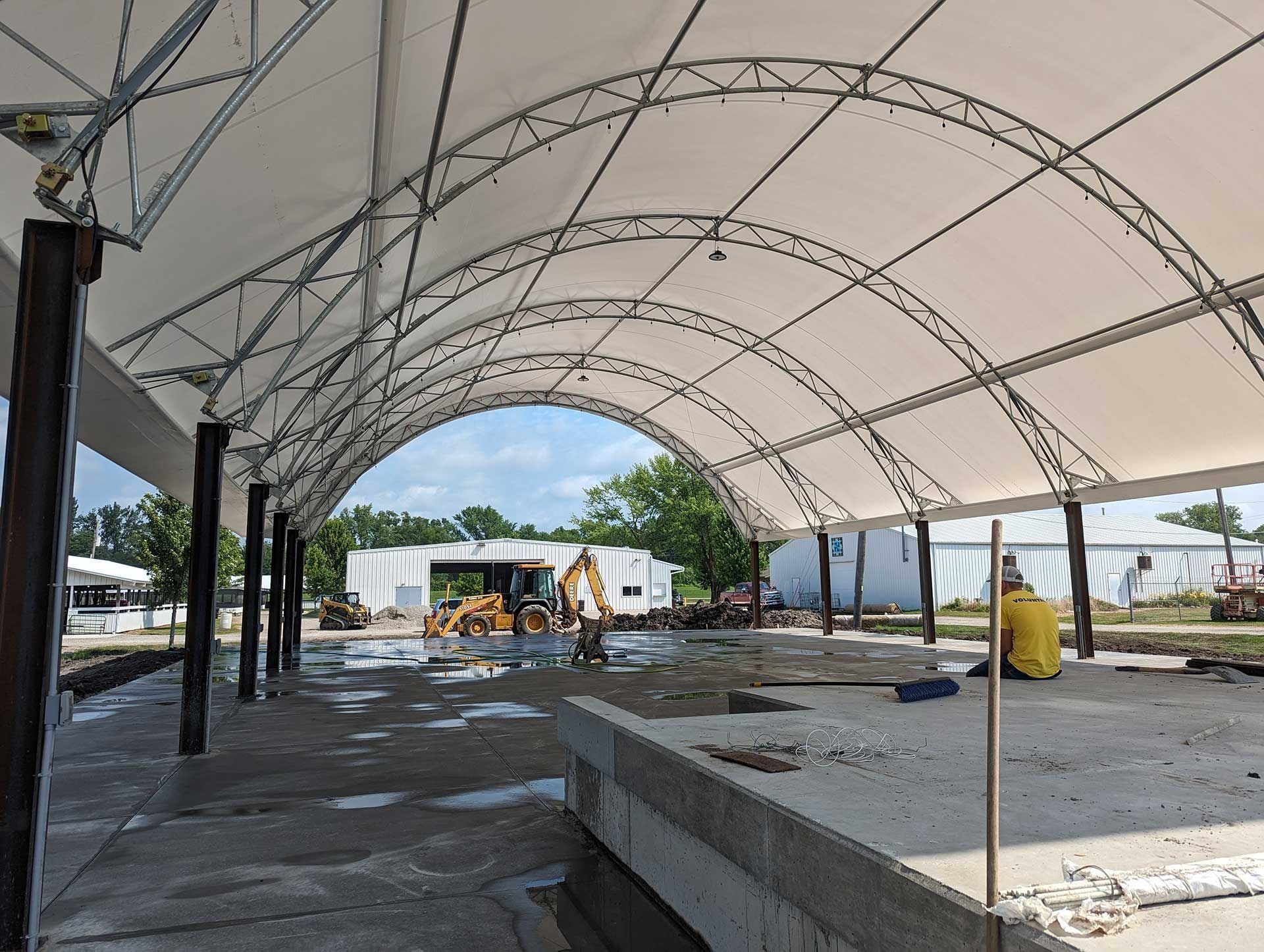 Construction site under a white canopy with arched metal supports. Workers and equipment visible.