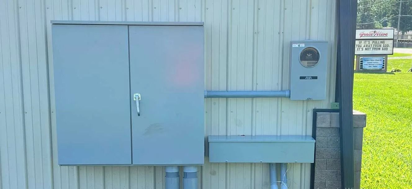 Electrical boxes and meter on a building's exterior with conduit and wiring. Green grass in background.