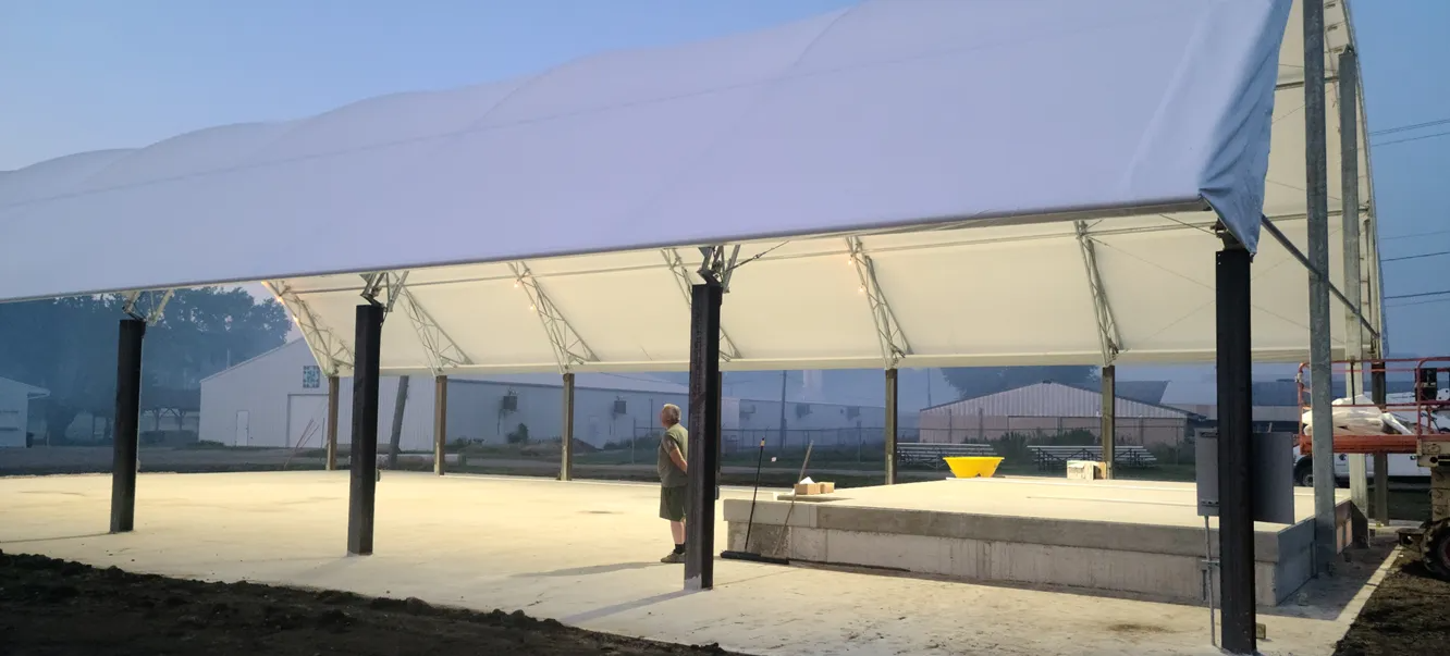 A large white tent with support beams on a concrete foundation in a field. A person stands inside the tent.