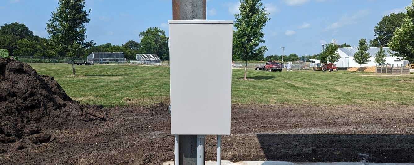 A white utility box attached to a pole in a grassy field with trees and buildings in the distance.