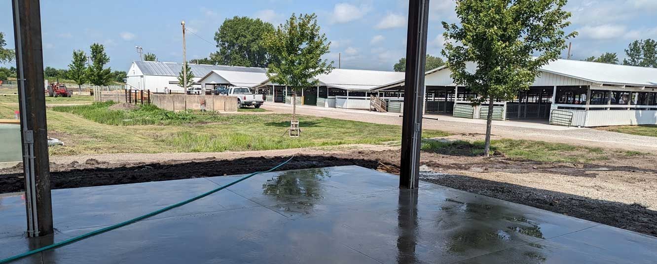 View from a dark, wet structure towards a farm with buildings, trees, and grass under a blue sky.