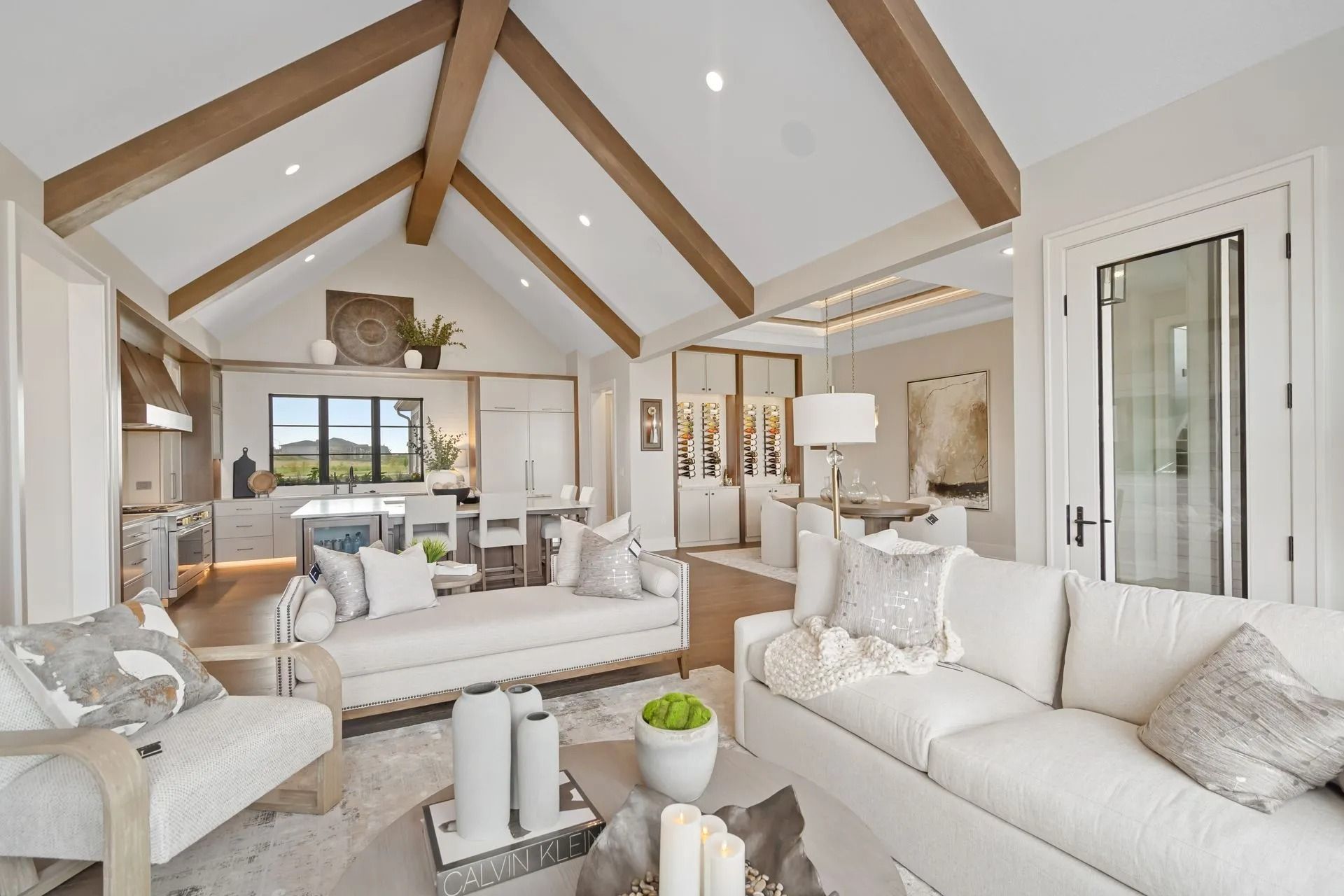 Living room with vaulted ceiling, exposed wood beams, white furniture, and an open kitchen.