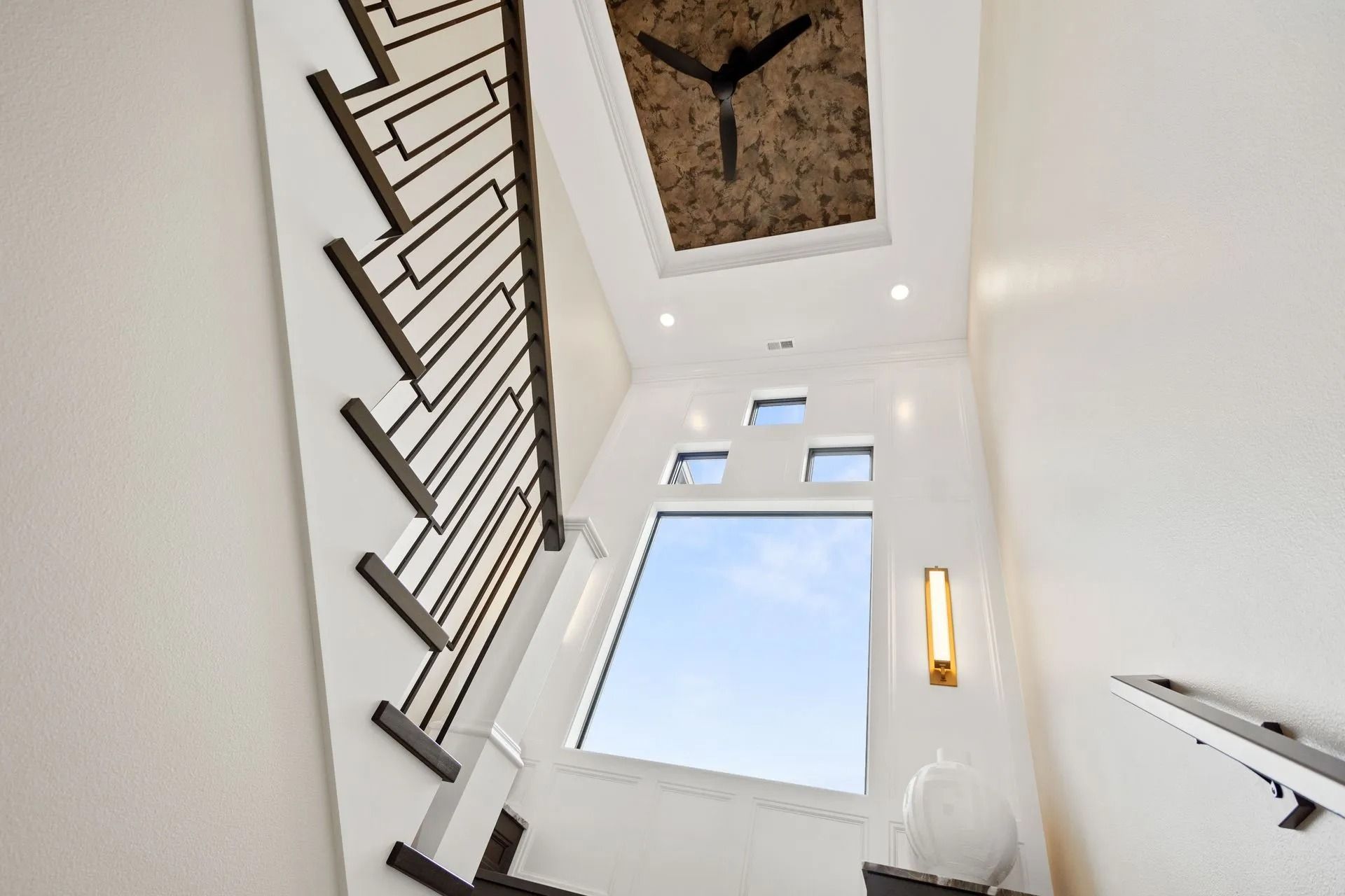 Staircase with geometric railing, large window, ceiling fan, and white walls.